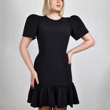 Load image into Gallery viewer, Close-up of the Femponiq Black Pleated Shoulder Peplum Hem Cady Dress in black, highlighting the pleated shoulder detail and luxurious fabric texture.  Showcasing a curvy woman, accentuating an hourglass shape with its bold silhouette.