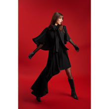 Load image into Gallery viewer, Model in the Femponiq Black Bow Tie Neck Cape Maxi Dress extending her arms to display the dramatic cape sleeves and flowing asymmetric hem, set against a striking red backdrop.
