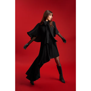Model in the Femponiq Black Bow Tie Neck Cape Maxi Dress extending her arms to display the dramatic cape sleeves and flowing asymmetric hem, set against a striking red backdrop.