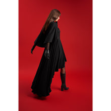 Load image into Gallery viewer, Model wearing the Femponiq Black Bow Tie Neck Cape Maxi Dress, walking in profile to showcase the dramatic cape back and flowing asymmetric hem against a vivid red backdrop.

