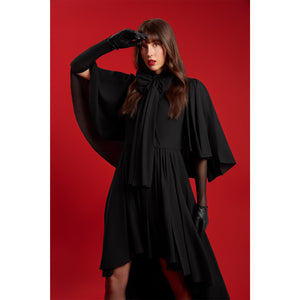 Model wearing the Femponiq Black Bow Tie Neck Cape Maxi Dress, featuring dramatic cape sleeves and a voluminous bow at the neckline, photographed against a bold red background.