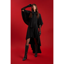 Load image into Gallery viewer, Model wearing the Femponiq Black Bow Tie Neck Cape Maxi Dress striking a dramatic pose with one arm raised, showcasing flowing cape sleeves and a high-low hemline, styled with black gloves and knee-high boots against a bold red background.
