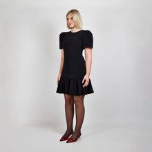 Load image into Gallery viewer, Side view of model in Femponiq Black Pleated Shoulder Peplum Hem Cady Dress, highlighting the dress’s sleek silhouette, pleated shoulder sleeves and feminine peplum accent.
