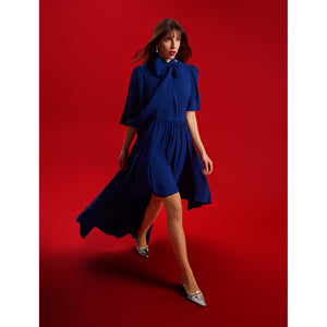 Model walking in Femponiq’s Midnight Blue Bow Tie Neck Cape Sleeve Maxi Dress, featuring a statement bow-tie neckline, dramatic cape sleeves, and an asymmetric hem with a shorter front and flowing maxi-length back. The voluminous drape and gathered waist create graceful movement against a deep red backdrop.