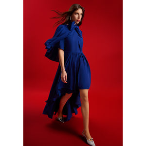 Model in motion wearing Femponiq’s Midnight Blue Bow Tie Neck Cape Sleeve Maxi Dress, showcasing the dramatic cape sleeves, gathered waist, and cascading high-low hem. The voluminous drape and flowing movement highlight the luxurious silhouette against a rich red background.