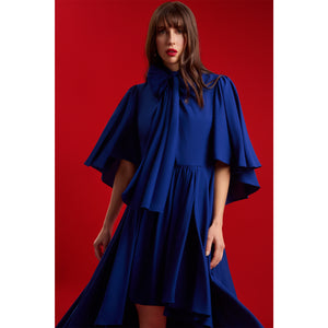 Close-up of a model wearing Femponiq’s Midnight Blue Bow Tie Neck Cape Sleeve Maxi Dress, highlighting the dramatic bow detail, cape sleeves and gathered waist against a bold red backdrop.