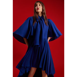 Close-up of a model wearing Femponiq’s Midnight Blue Bow Tie Neck Cape Sleeve Maxi Dress, showcasing the dramatic cape sleeves, oversized bow detail and cascading high–low skirt against a vivid red background.