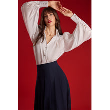 Load image into Gallery viewer, Model wearing the Femponiq High-Waisted Pleated Midi Skirt with asymmetric slits in navy, showing the flowing pleated silhouette and elegant movement against a red background.
