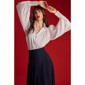 Model wearing the Femponiq High-Waisted Pleated Midi Skirt with asymmetric slits in navy, showing the flowing pleated silhouette and elegant movement against a red background.