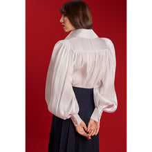 Load image into Gallery viewer, Model wearing the Femponiq Puff Sleeve V-Neck Gathered Shirt in sheer white, showing the gathered back yoke and voluminous sleeves against a red background.
