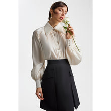 Load image into Gallery viewer, Close-up view of a model wearing the Femponiq pearl-white gathered-neck blouse with voluminous sleeves, paired with the Femponiq black pleated flared mini skirt. The model holds a white rose, highlighting the refined, feminine styling and the blouse’s subtle sheen and metallic buttons.
