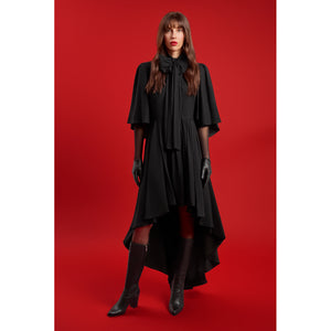 Model wearing the Femponiq Black Bow Tie Neck Cape Maxi Dress with dramatic cape sleeves, oversized bow detail, and high-low cascading hemline, styled with black gloves and knee-high boots against a red backdrop.