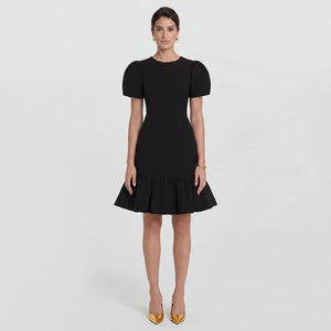 Brunette model wearing the Femponiq Black Pleated Shoulder Peplum Hem Cady Dress, highlighting the sophisticated pleated shoulder detail and the flattering, feminine peplum hem at full front view. The dress enhances an hourglass figure with its sculpted silhouette, offering a timeless and elegant design.