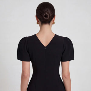 A close-up back view of model in Femponiq Black Pleated Shoulder Peplum Hem Cady Dress, emphasizing the structured fit and elegant V neck closure at the back.