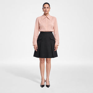 A model stands against a light gray studio background wearing Femponiq's dusty rose pink long-sleeve collared shirt and black pleated A-line skirt with flap pockets, paired with black pointed heels. Front Look