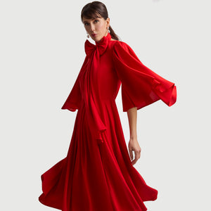 Side view of the Femponiq Gathered Midi Red Bow Tie Neck Dress, showing the dramatic full-circle sleeves, statement bow neckline, and flowing pleated skirt. The soft, draped fabric enhances movement and creates an elegant, feminine silhouette.