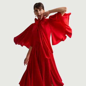 Close-up view of the Femponiq Gathered Midi Red Bow Tie Neck Dress, highlighting the dramatic full-circle sleeves, elegant bow neckline, and flowing pleated fabric. The fluid drape and side slits enhance movement, creating a graceful and sophisticated silhouette.