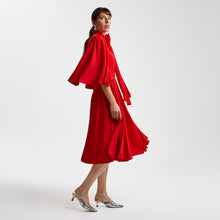 Load image into Gallery viewer, Right-side view of the Femponiq Gathered Midi Red Bow Tie Neck Dress, showcasing the flowing full-circle sleeves, statement bow neckline, and mid-length pleated skirt with front slits. The fluid fabric enhances movement and creates an elegant, feminine silhouette.
