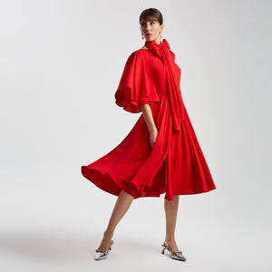 Side view of the Femponiq Gathered Midi Red Bow Tie Neck Dress, showing the dramatic full-circle sleeves, flowing pleated skirt, and front slits. The bow neckline and fluid movement highlight the dress’s graceful and sophisticated silhouette.