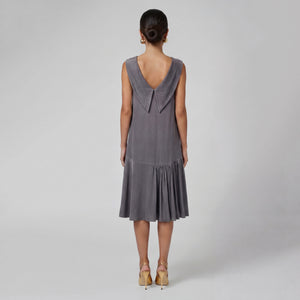 Woman facing backwards wearing a soft gray sleeveless midi dress with a draped neckline, relaxed silhouette, and asymmetrical gathered hem. Styled with gold pointed heels and gold statement earrings against a light gray background. Elegant modern dress by Femponiq.