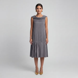Woman facing front wearing a soft gray sleeveless midi dress with a draped neckline, relaxed silhouette, and asymmetrical gathered hem. Styled with gold pointed heels and gold statement earrings against a light gray background. Elegant modern dress by Femponiq.