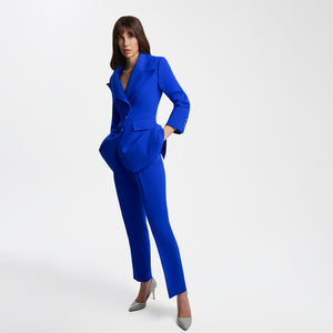 Full-length view of a model wearing bright blue high-waisted tailored trousers with pressed front pleats, paired with a matching structured blazer. The Femponiq trousers feature a sleek long-leg silhouette, functional belt, concealed snap fastening, and slash pockets, creating a polished and modern look.