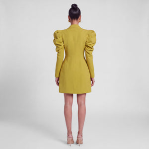A woman viewed from the back wearing the Femponiq mustard yellow hourglass blazer dress with dramatic puffed long sleeves and structured shoulders, paired with nude high-heeled sandals against a light gray studio background.
