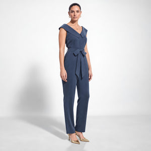 Model wearing Femponiq’s denim blue vegan cupro jumpsuit with a folded off-shoulder collar, self-tie waist belt and tapered legs, three-quarter side view.