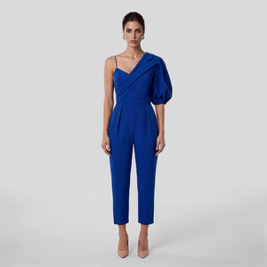 A model stands against a light gray studio background wearing a royal blue one-shoulder jumpsuit with a puffed sleeve, thin spaghetti straps, a wrap-style bodice, and cropped straight-leg pants