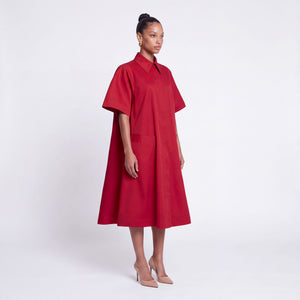 A model stands in profile wearing a bright red midi dress with a high collar, short wide sleeves, and front patch pockets, paired with nude high heels, against a plain light gray studio background.