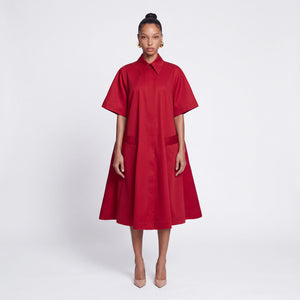 A model stands against a light gray studio background wearing a bright red midi dress with short sleeves, a pointed collar, and large front patch pockets.