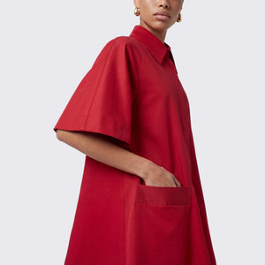 A model wearing a red oversized collared shirt dress with short sleeves and a front pocket, shown from the side against a light gray studio background.