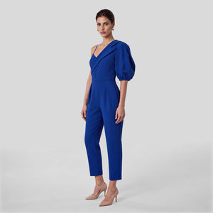 A woman modeling a royal blue one-shoulder jumpsuit with puff sleeves and slim cropped legs, paired with nude high heels, against a light gray studio background.