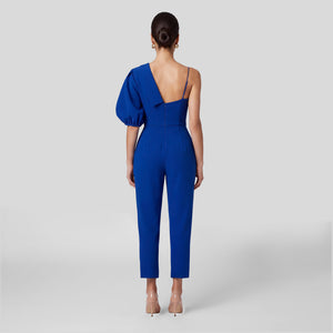 A woman viewed from the back wearing a royal blue one-shoulder jumpsuit with a structured puff sleeve, thin straps, and straight cropped legs, paired with clear high heels against a light gray studio background.