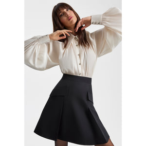 Close-up view of a model wearing a pearl-white gathered-neck shirt with voluminous raglan sleeves and metallic buttons, paired with a black tailored mini skirt. The shirt’s semi-sheer fabric creates soft movement and a refined, elegant look.