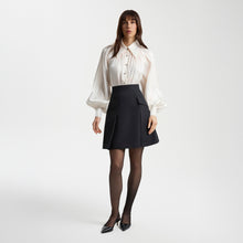Load image into Gallery viewer, Model wearing a pearl-white gathered-neck shirt with voluminous raglan sleeves paired with a black tailored mini skirt. The semi-sheer shirt subtly sparkles and features metallic buttons, creating a feminine, elegant look.
