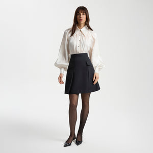 Model wearing a pearl-white gathered-neck shirt with voluminous raglan sleeves paired with a black tailored mini skirt. The semi-sheer shirt subtly sparkles and features metallic buttons, creating a feminine, elegant look.