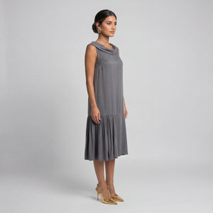 Woman facing side wearing a soft gray sleeveless midi dress with a draped neckline, relaxed silhouette, and asymmetrical gathered hem. Styled with gold pointed heels and gold statement earrings against a light gray background. Elegant modern dress by Femponiq.