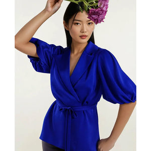 Woman wearing a vibrant cobalt blue wrap-style blouse with voluminous puff sleeves, a tailored lapel collar, and a self-tie waist belt. Styled with sleek hair and floral accents for an elegant, statement look by Femponiq.