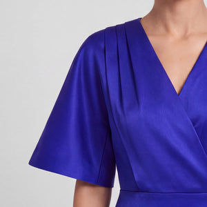 Close-up of a model wearing a royal blue wrap-style dress with short flared sleeves and a V-neckline, shown against a light gray studio background.