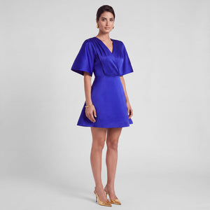 A woman modeling a royal blue satin mini dress with short flutter sleeves, a V-neckline, and a flared skirt, paired with gold heels against a light gray studio background. Side Look