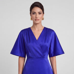 A woman wearing a royal blue satin wrap dress with short flared sleeves and a V-neckline, photographed against a light gray studio background.