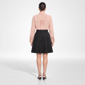 A model viewed from the back wearing Femponiq's dusty rose pink long-sleeve tailored shirt with pleated detailing on the upper back and a black A-line skirt, paired with black heels, against a light gray studio background.