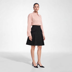 A model stands against a light gray studio background wearing Femponiq's dusty rose pink long-sleeve button-up tailored shirt, black pleated A-line skirt with flap pockets, and black pointed-toe heels.