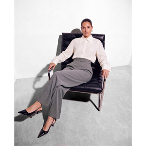 
Model leaning back in a black leather chair wearing a Femponiq ivory pointed-collar shirt with wide cuffs, grey high-waisted tailored trousers and black pointed-toe slingback heels.
