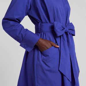A close-up of a person wearing a royal blue trench coat with a matching waist tie and large front pockets, shown against a plain light background.