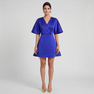 A woman modeling a royal blue satin mini dress with short flutter sleeves, a V-neckline, and a flared skirt, paired with gold heels against a light gray studio background. Front Look