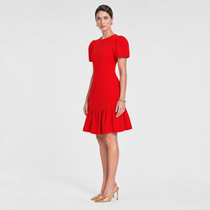 Woman standing side wearing a bold red fitted midi dress with structured puff sleeves, a cinched waist, and a flared ruffle hem. Styled with gold pointed heels and minimal gold jewelry against a light gray background. Elegant feminine silhouette by Femponiq.