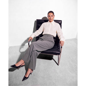 
Model leaning back in a black leather chair wearing a Femponiq ivory pointed-collar shirt with wide cuffs, grey high-waisted tailored trousers and black pointed-toe slingback heels.