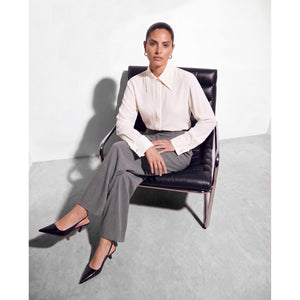
Model seated in a black leather chair wearing a Femponiq ivory pointed-collar shirt with wide cuffs, grey high-waisted tailored trousers and black pointed-toe slingback heels.